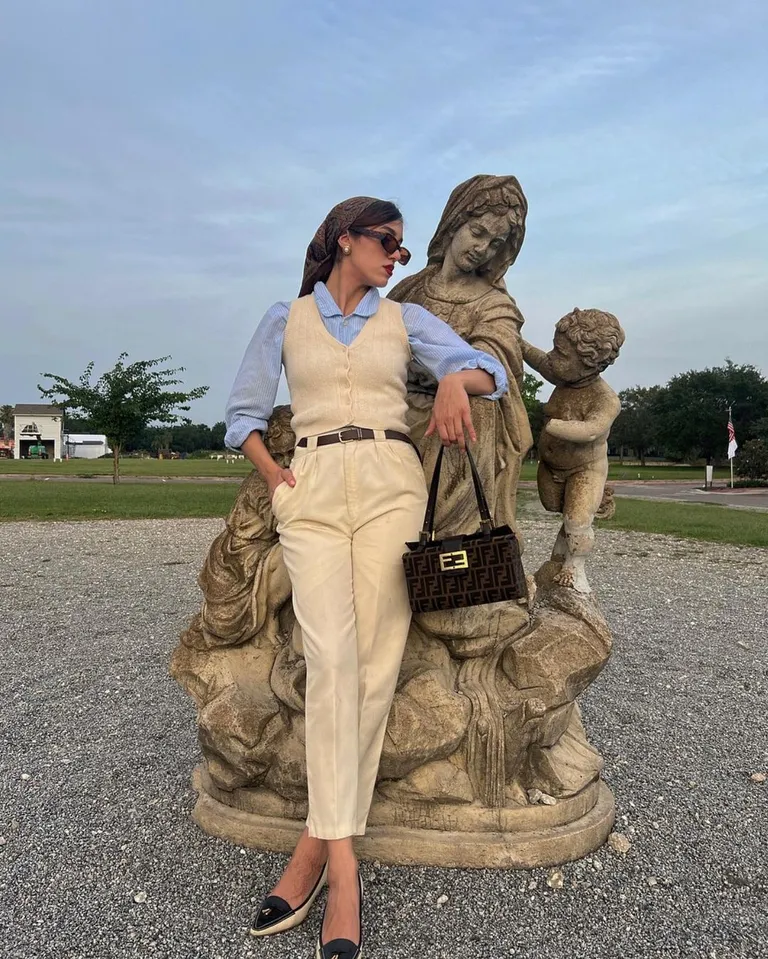 Woman in beige tailored trousers, a blue shirt, and a beige vest with a headscarf, styled as a chic and polished modest summer outfit.