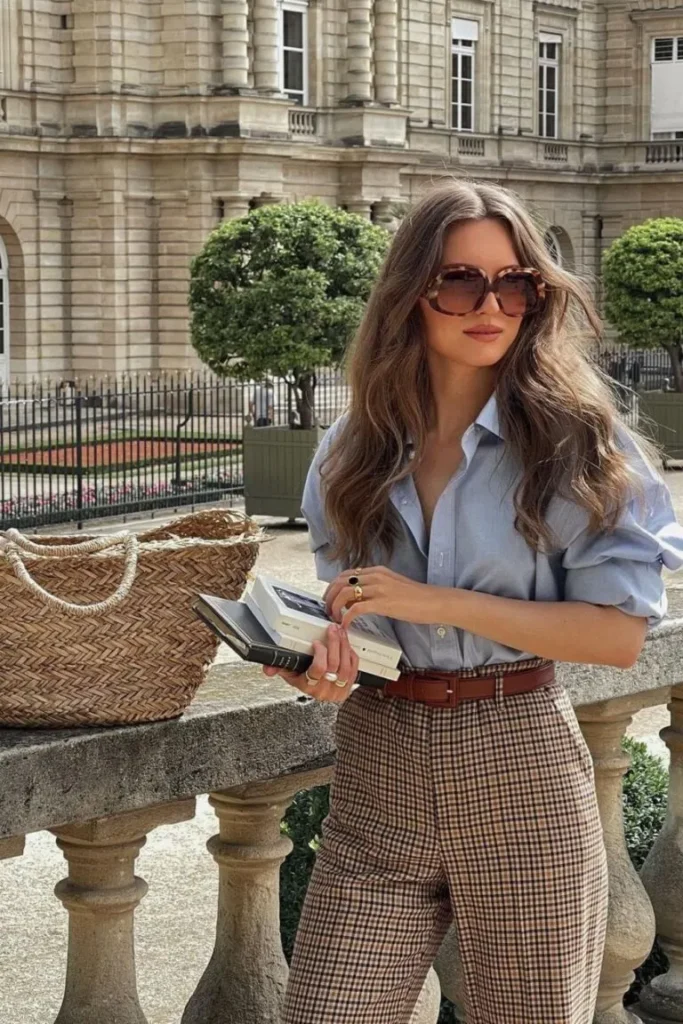 Woman in light blue shirt tucked into checkered high-waisted trousers, holding books and wearing sunglasses for a look expensive outfit.