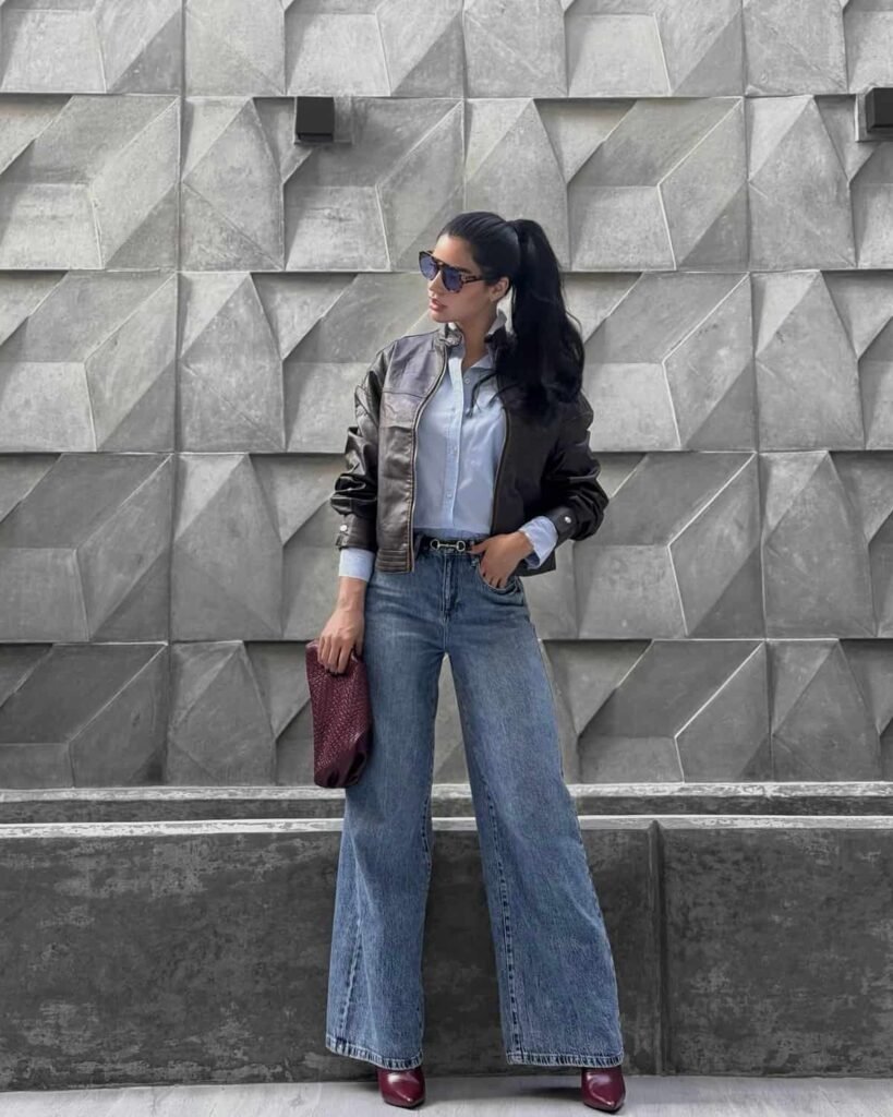 Bold wide leg pants outfit combining mid-wash jeans, a tucked-in blue button-up shirt, and a brown leather bomber jacket.