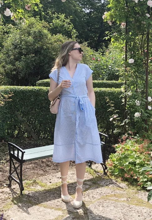 A woman in a light blue belted dress with espadrille wedges, standing in a rose garden – romantic and timeless old money spring style.