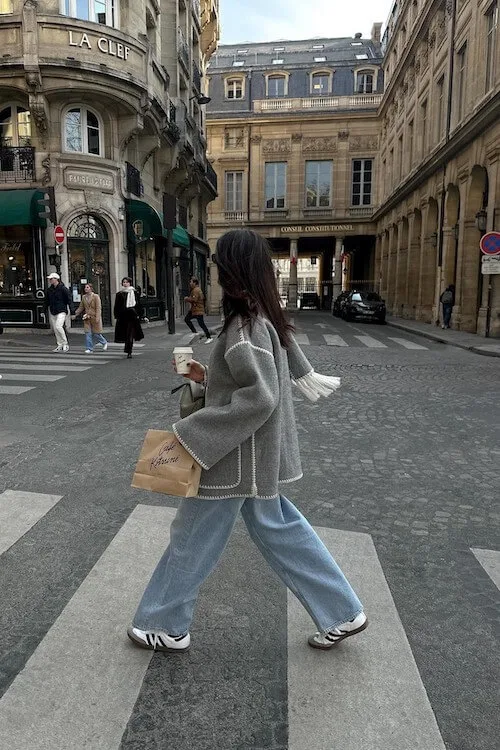 Parisian-inspired casual look with Adidas Samba Outfits, flared light-wash jeans, grey wool coat, and shopping bag.