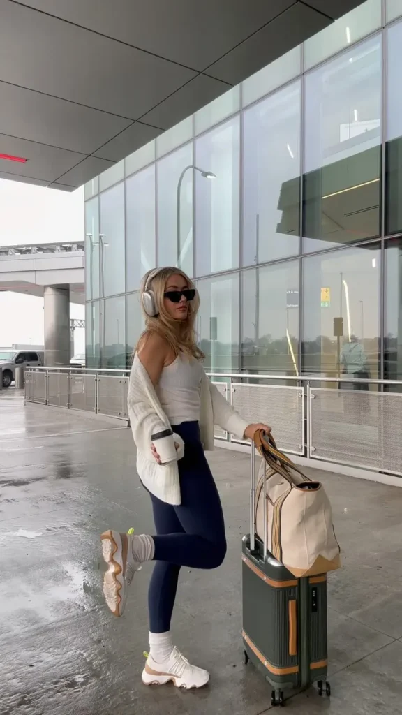 Cute Airport Outfits: Woman in navy leggings, white tube top, and cozy cardigan, wearing large headphones and white sneakers, leaning on a green and tan suitcase.