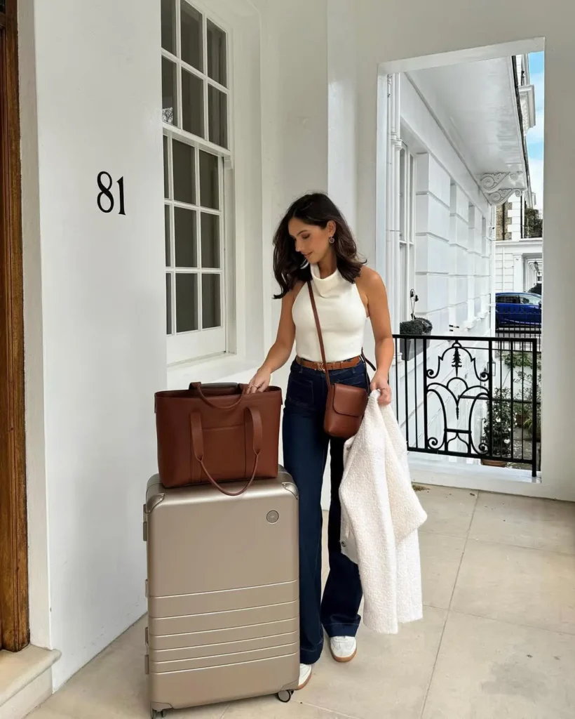 Cute Airport Outfits: Sophisticated look with a sleeveless white top, high-waisted flared jeans, and brown leather bag, paired with a champagne-gold suitcase.