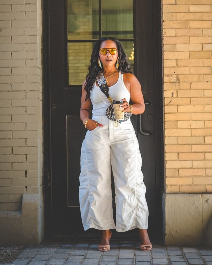 Bold and confident Summer Outfits look with a white tank top and wide-leg cream trousers, accessorized with yellow-tinted sunglasses and gold jewelry.
