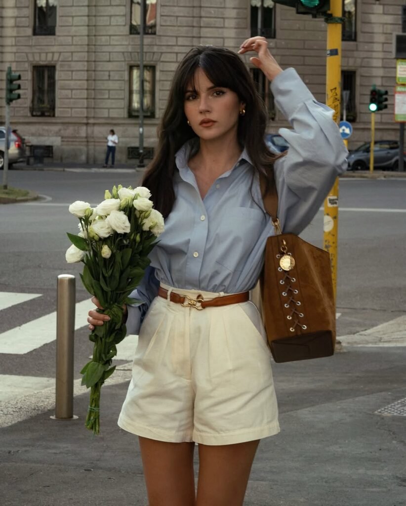 Woman in a sky blue oversized shirt tucked into ivory pleated shorts, holding white flowers for a soft look expensive outfit.