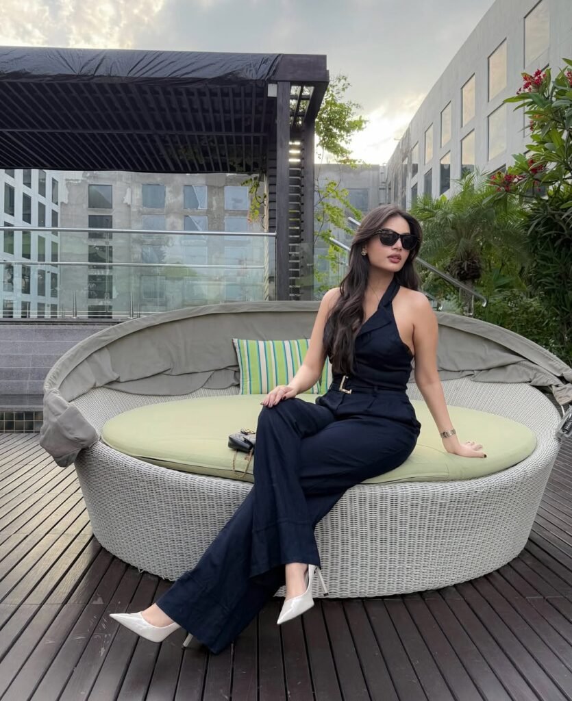 A woman in a chic black halter-neck jumpsuit with a gold-buckled belt, white heels, and sunglasses, sitting on a rooftop lounge – modern old money spring outfit with refined flair.