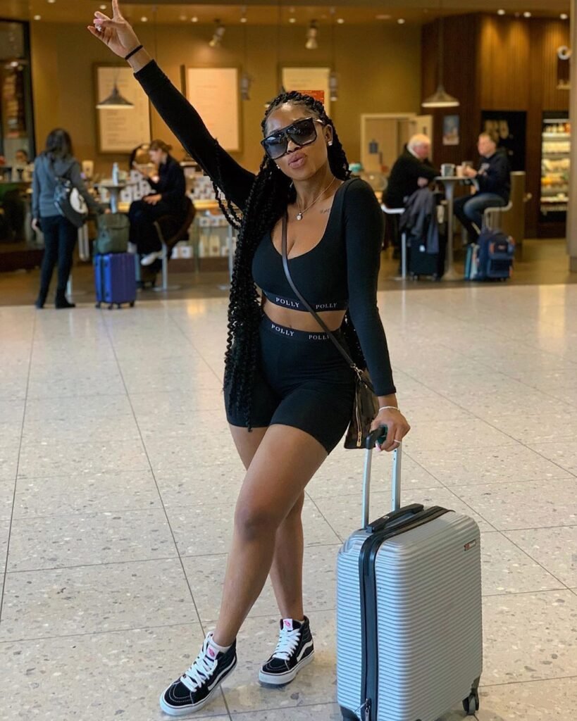 Cute Airport Outfits: Woman in black two-piece sporty set with long braids and sunglasses, posing confidently with a silver carry-on suitcase.
