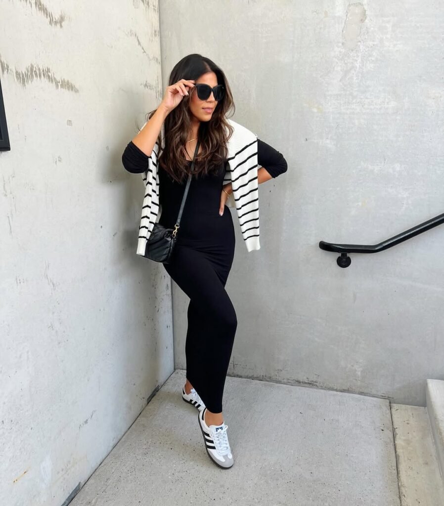 Chic Adidas Samba Outfits styled with a black fitted dress, a striped sweater casually thrown over the shoulders, oversized sunglasses, and white Samba sneakers.