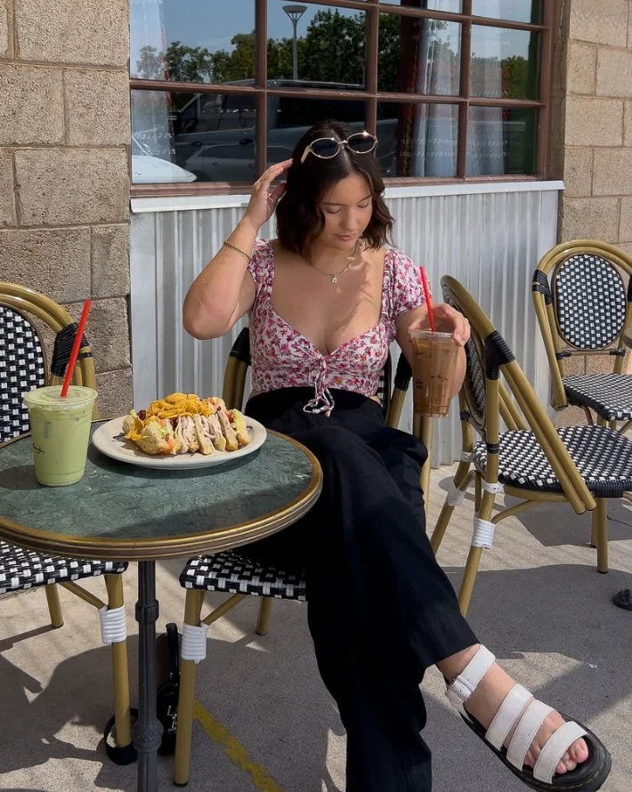 Coffee date outfit featuring a floral top with a tie-front design, paired with black pants and white sandals, enjoying iced drinks and snacks at a café.