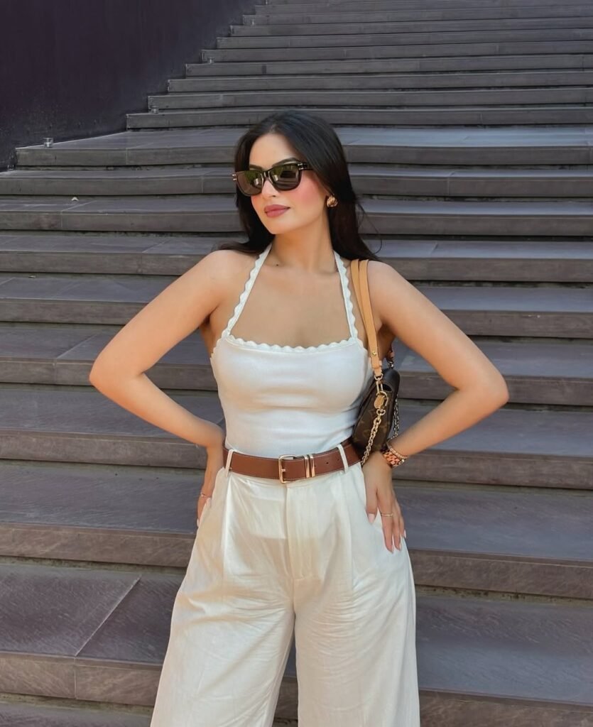 A woman dressed in a white scallop-trimmed top and high-waisted cream trousers with a brown belt, accessorized with dark sunglasses – effortless old money spring elegance.
