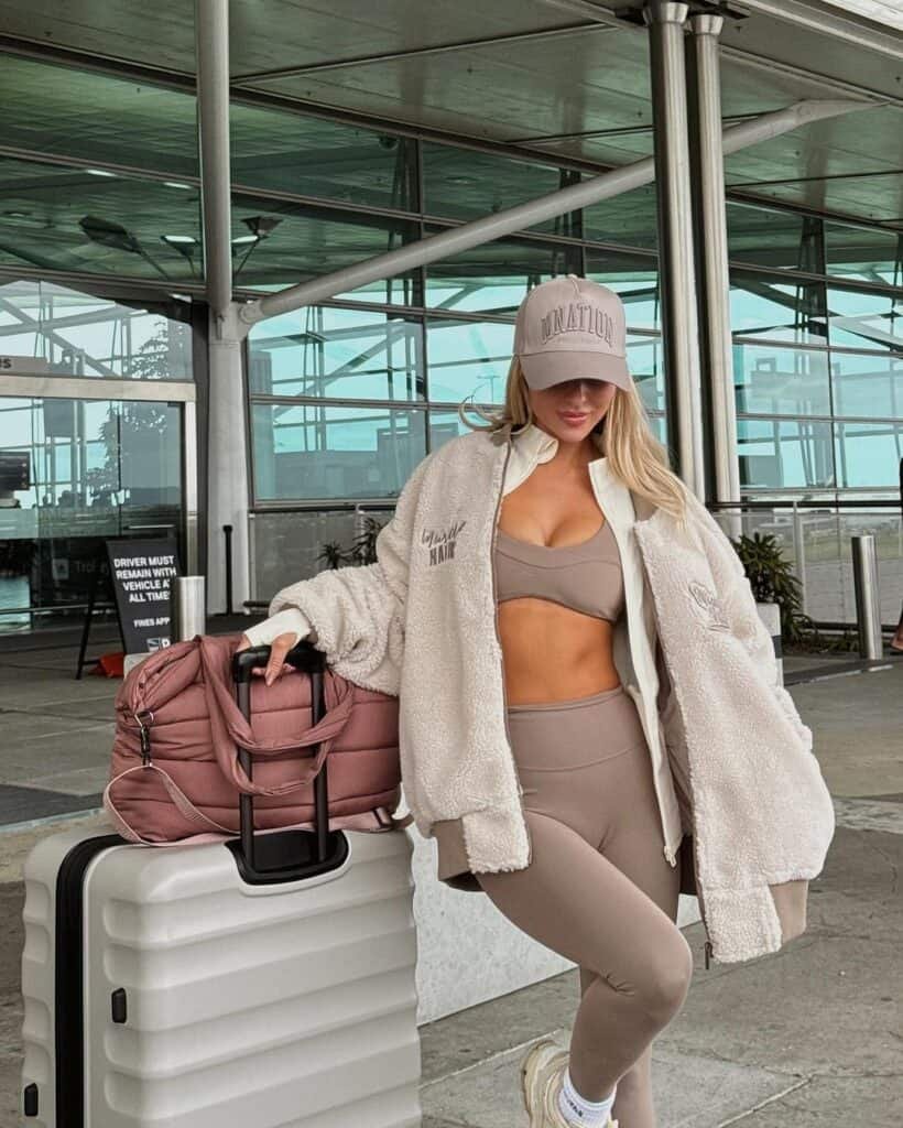 Cute Airport Outfits: Woman in taupe sports bra and leggings with a teddy coat and cap, leaning on a white suitcase with a blush duffel bag.