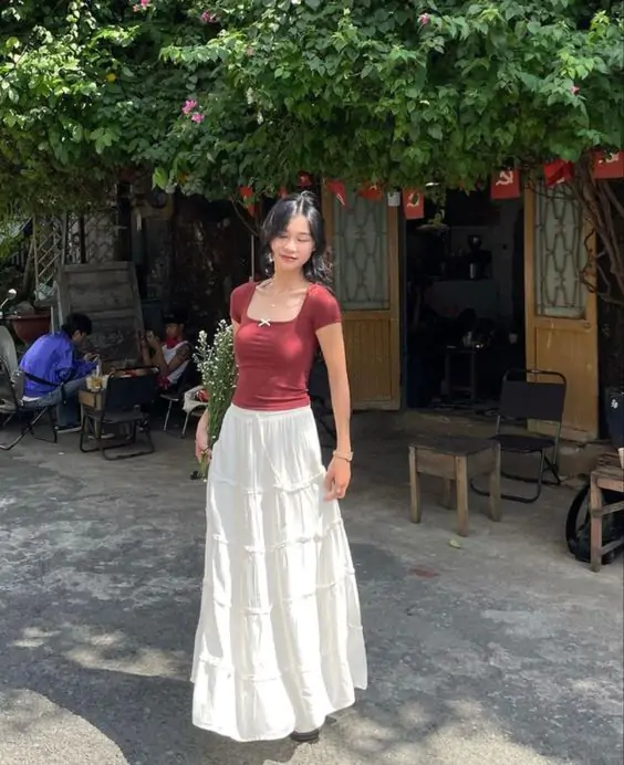 Bohemian-inspired Summer Outfits look with a maroon fitted top and flowing white maxi skirt, standing gracefully in a sunlit outdoor café area.