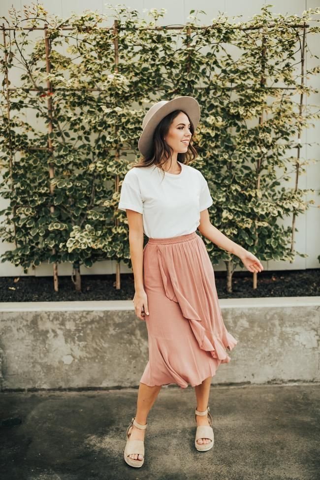 Woman in a flowy blush-pink midi skirt, white tee, and a sun hat, embodying a soft, feminine modest summer outfit.