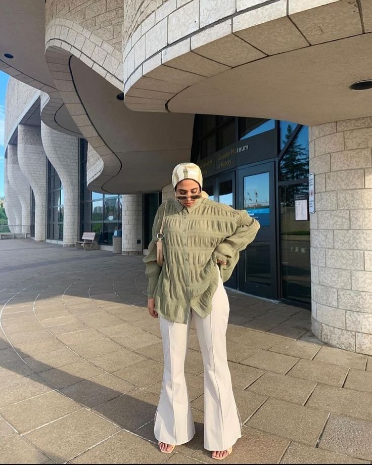 Woman in white flared pants and a loose, olive-green blouse with a hijab, styled modestly for a summer day out near an architectural landmark.