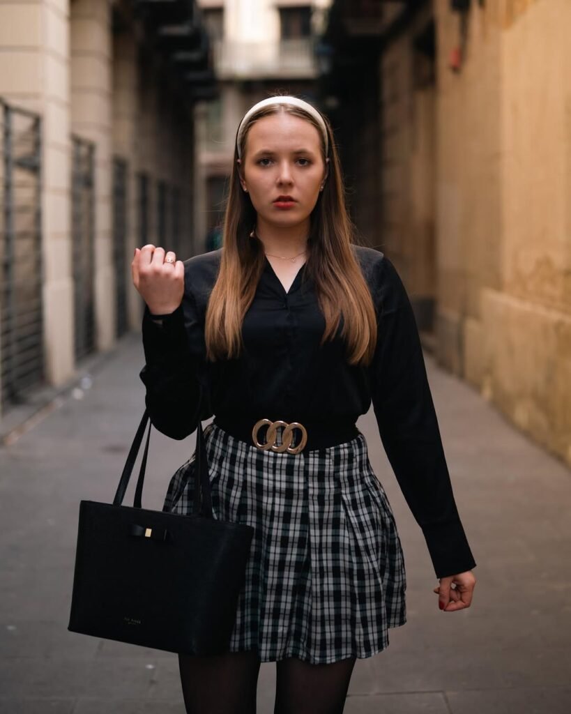 A woman wearing a black blouse tucked into a black-and-white plaid pleated skirt, black tights, and a white headband, standing in a European alley – timeless old money spring school-girl look.