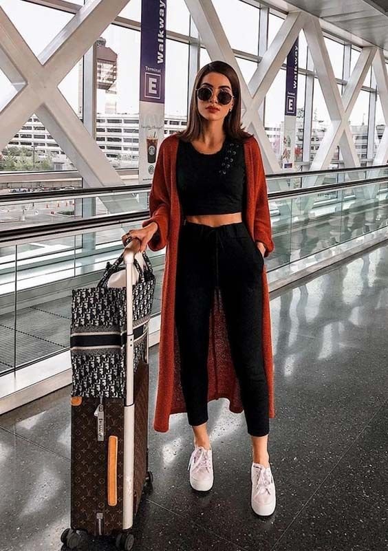 Cute Airport Outfits: Woman in black crop top and joggers, layered with a rust-colored duster coat, posing with a Louis Vuitton suitcase at an airport terminal.