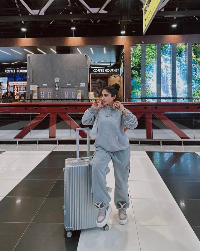 Cute Airport Outfits: Casual style with an oversized grey hoodie and joggers, pink-accent sneakers, and a silver ribbed suitcase, inside a modern airport.