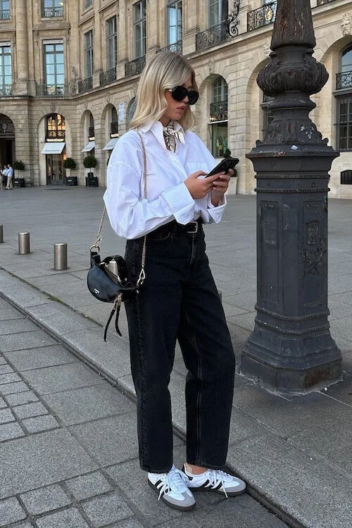 Parisian-inspired Adidas Samba Outfits with a white oversized shirt tucked into high-waisted black jeans, accessorized with a mini bag, silk scarf, and Samba sneakers.