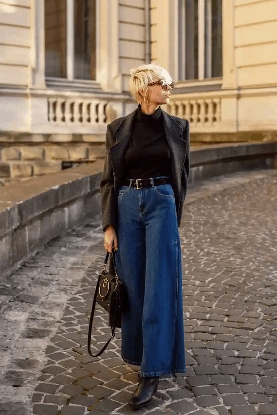 Vintage-inspired wide leg pants outfit combining a black turtleneck, charcoal blazer, and dark blue jeans with boots.