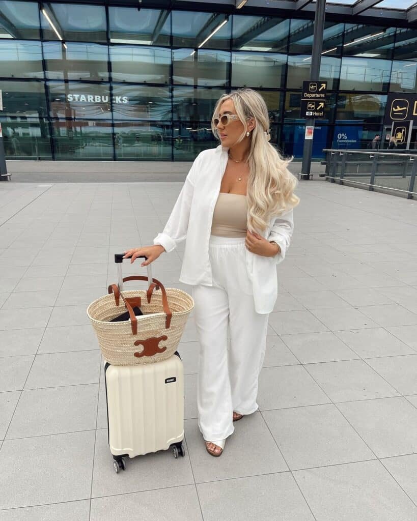 Cute Airport Outfits: Woman in an all-white set with flowy pants, tube top, and an open shirt, accessorized with sunglasses and a straw tote atop a cream suitcase.