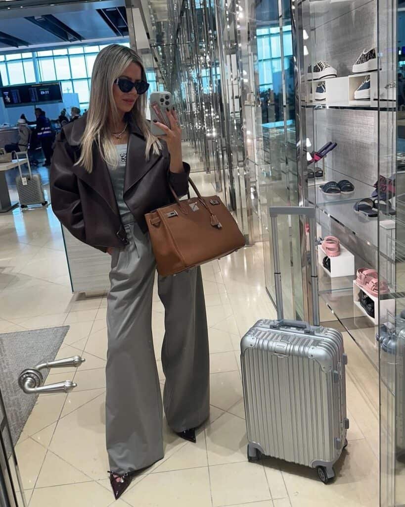 Cute Airport Outfits: Stylish traveler in grey wide-leg pants, white top, and oversized leather jacket, taking a mirror selfie with a silver suitcase and brown handbag.