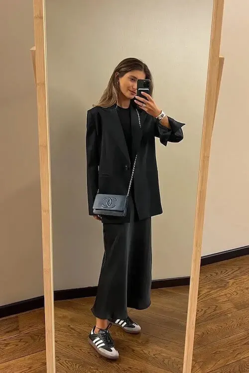 Monochrome Adidas Samba Outfits with an all-black ensemble featuring a long black skirt and oversized blazer, styled with a navy Chanel bag and black Samba sneakers.

