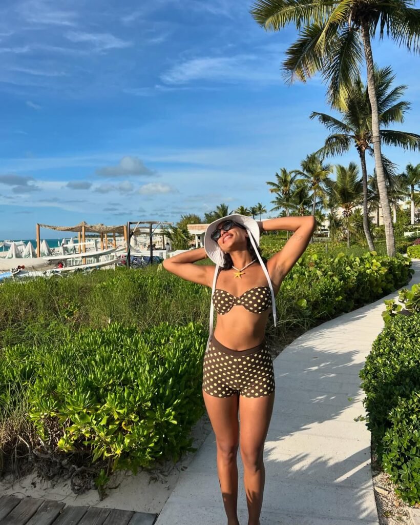 Retro-chic Swim Shorts outfit with a brown high-waisted polka dot set, paired with a matching bandeau top and white sun hat on a tropical boardwalk.
