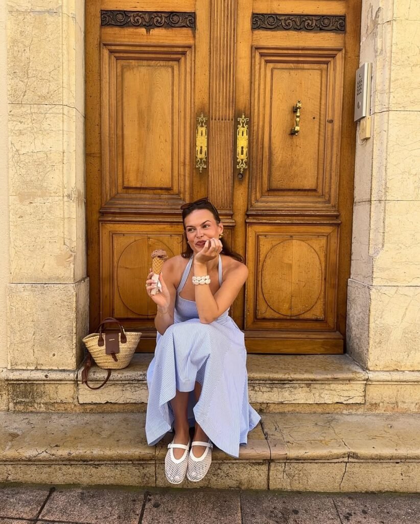 European Summer Outfit: Light blue halter-neck maxi dress with subtle stripes, white woven shoes, and a straw bag, sitting on stone steps enjoying ice cream.