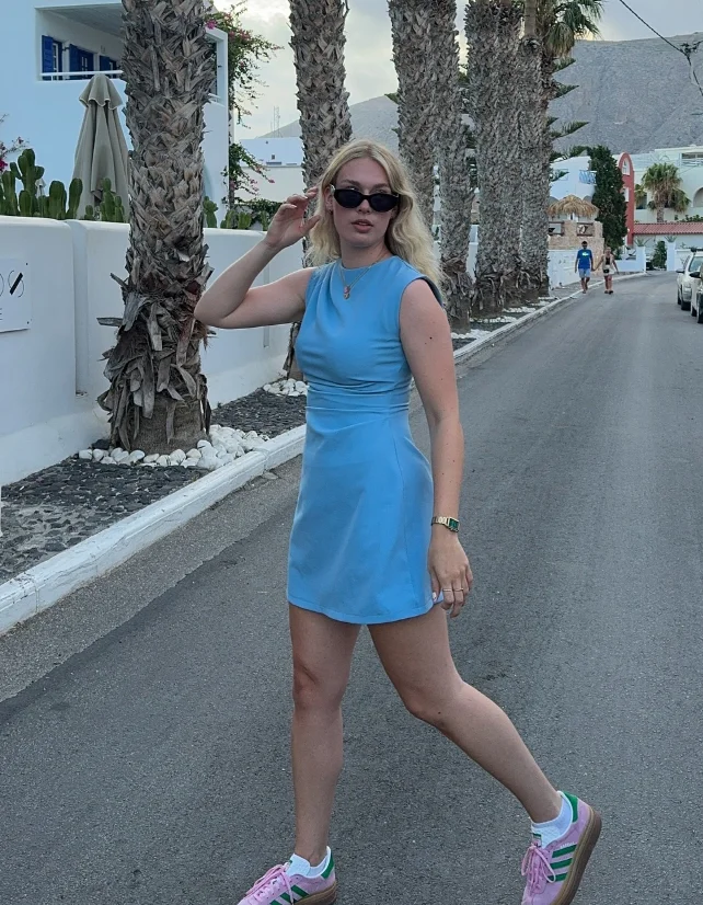 A woman walking on a palm-lined street wearing a sleeveless sky blue mini dress with pink and green Adidas sneakers; a perfect example of cute simple outfits for summer strolls.