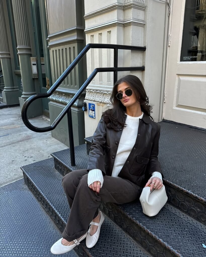 Woman in chill outfit ideas wearing a dark brown leather jacket, white sweater, brown pants, and white Mary Jane flats, holding a white handbag while sitting on metal steps.
