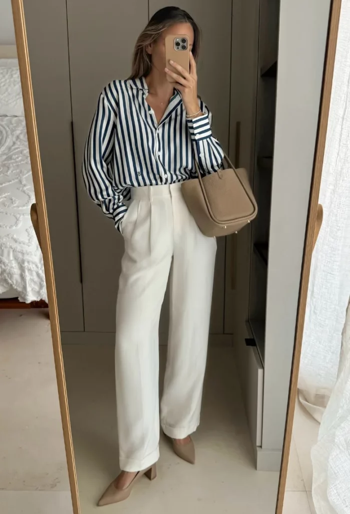 Office outfit featuring a blue-and-white striped button-down shirt tucked into high-waisted white tailored trousers, styled with beige pointed-toe heels and a taupe structured handbag.