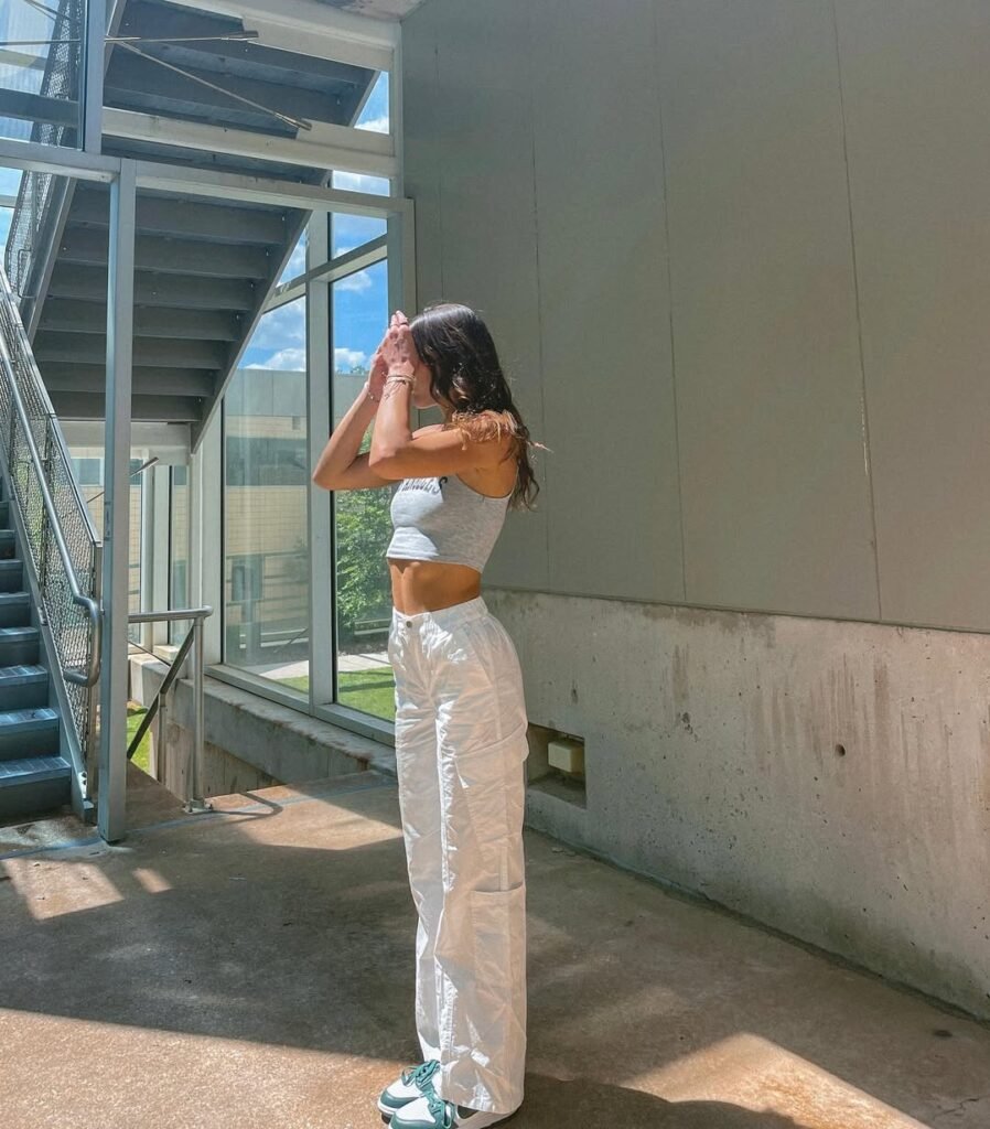 Woman wearing White Pants Outfits with a gray crop top, loose-fit cargo-style white pants, and teal sneakers, posing in a sunlit modern building.