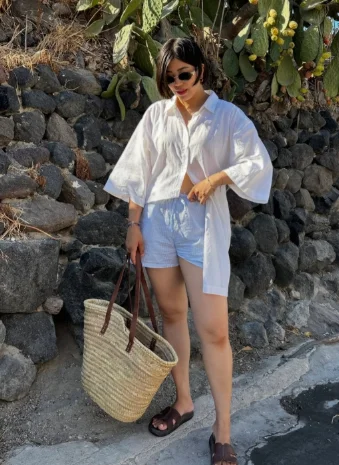 European Summer Outfit: Loose white button-up shirt with light blue shorts, brown sandals, and a large straw tote bag, standing by a rocky coastal road.