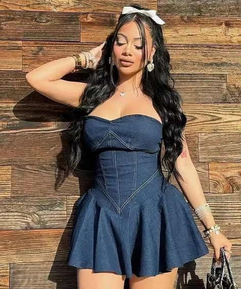 Woman in casual summer dress made of denim with a strapless corset top and flared mini skirt, accessorized with bracelets and a hair bow, posing outdoors in sunlight.