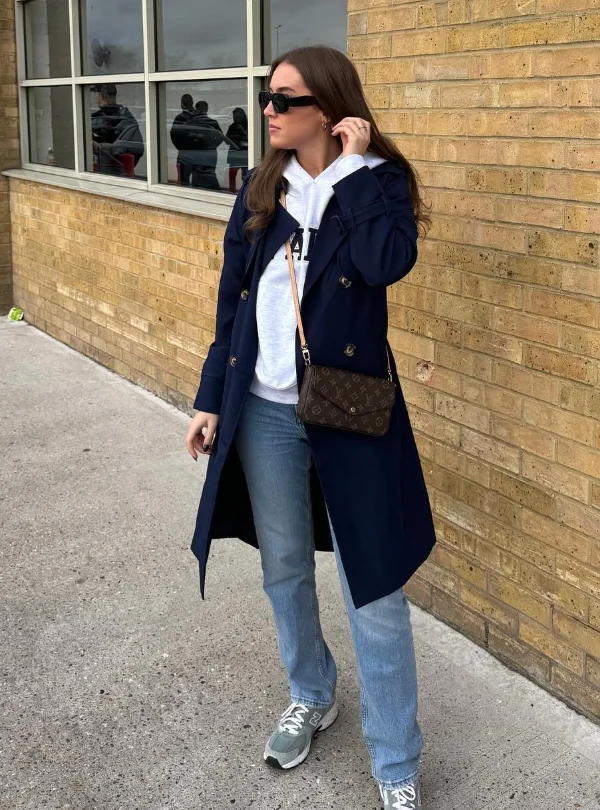 Woman in chill outfit ideas wearing a navy trench coat over a white hoodie, light blue jeans, grey sneakers, and a small Louis Vuitton crossbody bag.