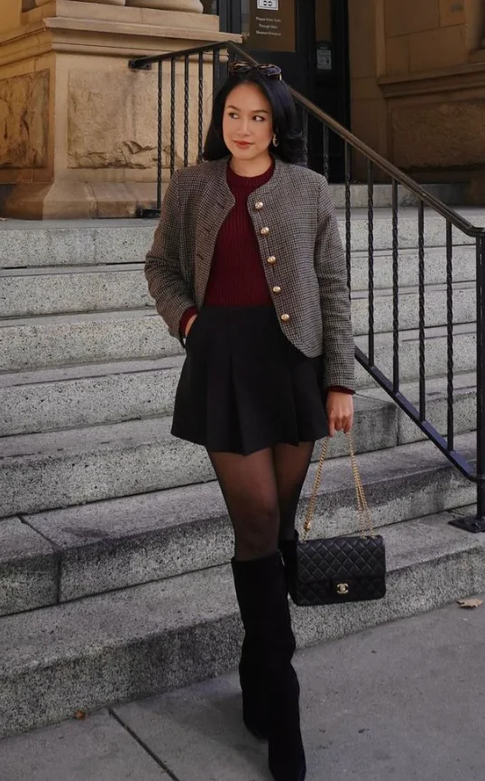 Woman in burgundy outfit wearing a burgundy sweater, black mini skirt, houndstooth jacket, black tights, and knee-high boots, holding a quilted handbag.