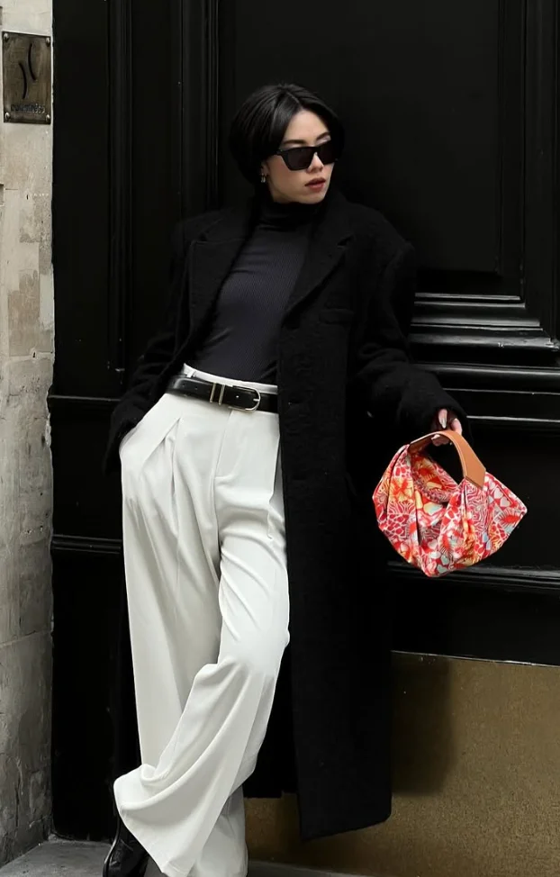 Stylish White Pants Outfits with a black coat, black turtleneck, and pleated wide-leg white trousers, accessorized with sunglasses and a colorful printed handbag.