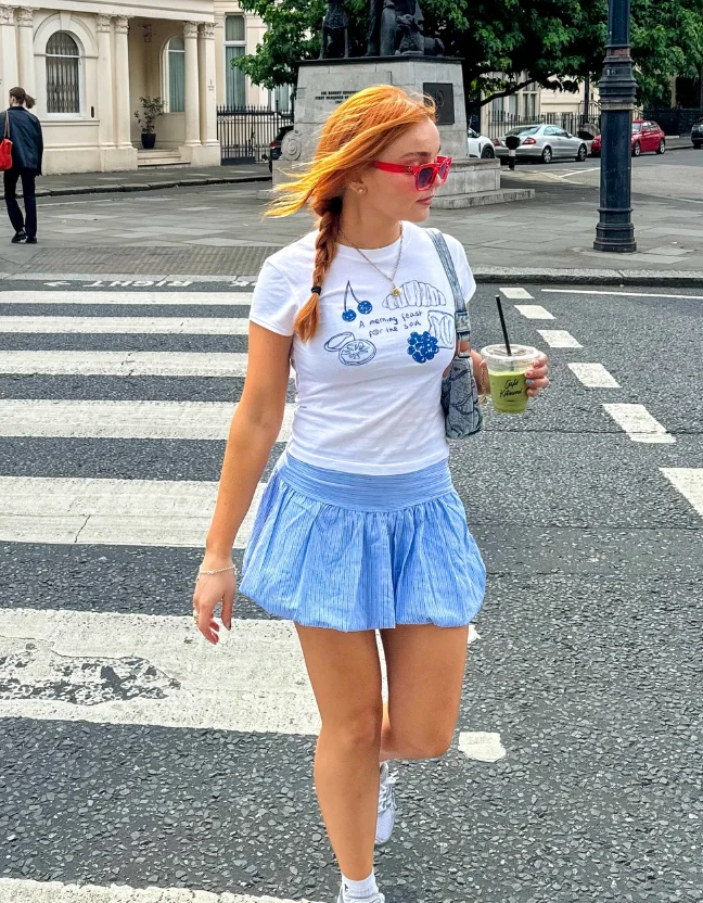 A woman crossing the street in a graphic tee and a light blue pleated mini skirt, accessorized with pink sunglasses and white sneakers – a youthful example of cute simple outfits.