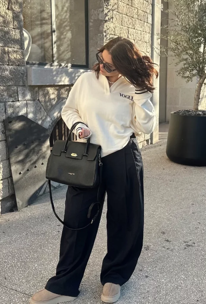 Woman in chill outfit ideas with a cream long-sleeve “Vogue” top, wide-leg black trousers, beige slip-on shoes, and a structured black handbag.