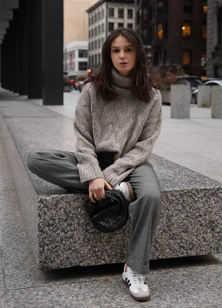 Casual grey trouser outfit with a chunky beige turtleneck sweater, black woven clutch bag, and white sneakers, styled on a granite bench in the city.
