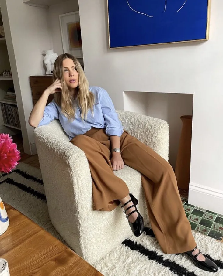 **Brown Trousers Outfits** – Woman sitting on a cream boucle armchair in a cozy living room, wearing wide-leg camel brown trousers, a light blue button-up shirt, and black patent Mary Jane flats, with long blonde hair styled in loose waves.
