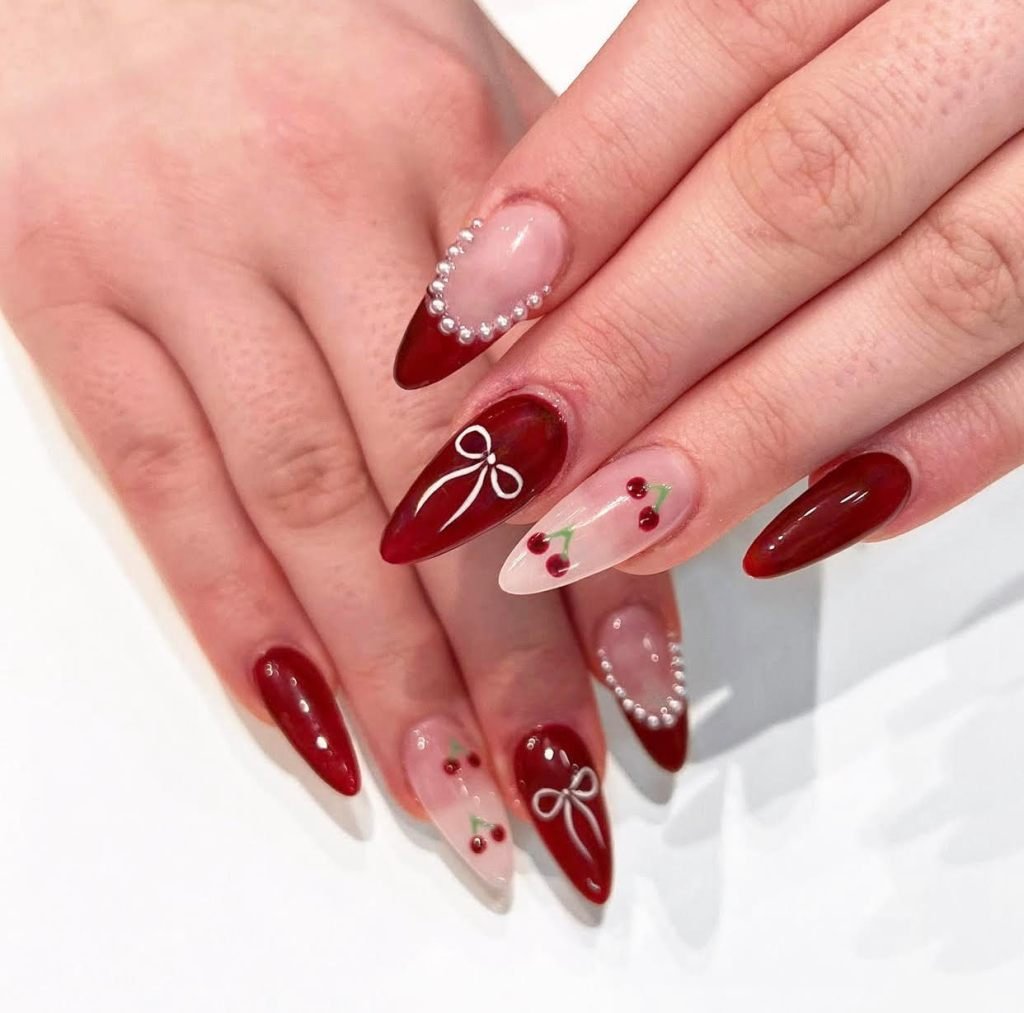 Long almond nails with deep red polish, pearl accents, white bows, and cherry nail art on nude bases.