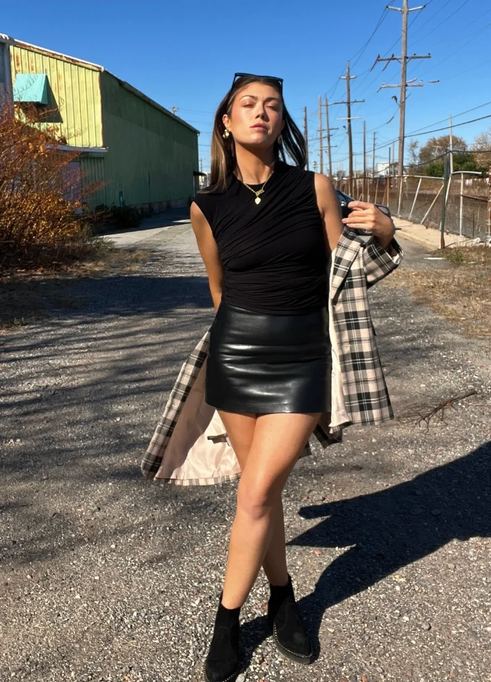 A cute simple outfit featuring a black sleeveless top, black leather mini skirt, and black ankle boots, styled with a plaid coat for an edgy chic vibe.