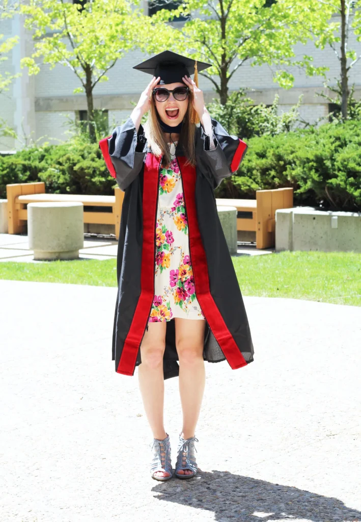 graduation outfit ideas: Short multicolored floral dress under a black graduation gown with red trim, paired with a graduation cap and oversized sunglasses.