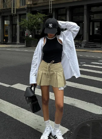 European Summer Outfit featuring a sporty chic look with a black sleeveless top, khaki pleated mini skirt, oversized white shirt, NY cap, and sneakers.