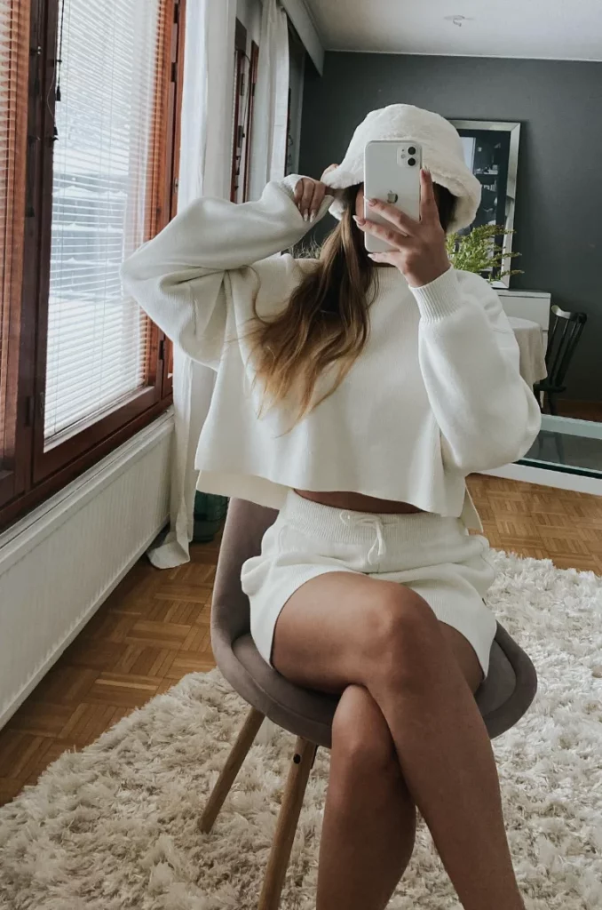 Woman in chill outfit ideas wearing a cropped white sweater, matching white knit shorts, and a white bucket hat, taking a mirror selfie while sitting on a chair indoors.