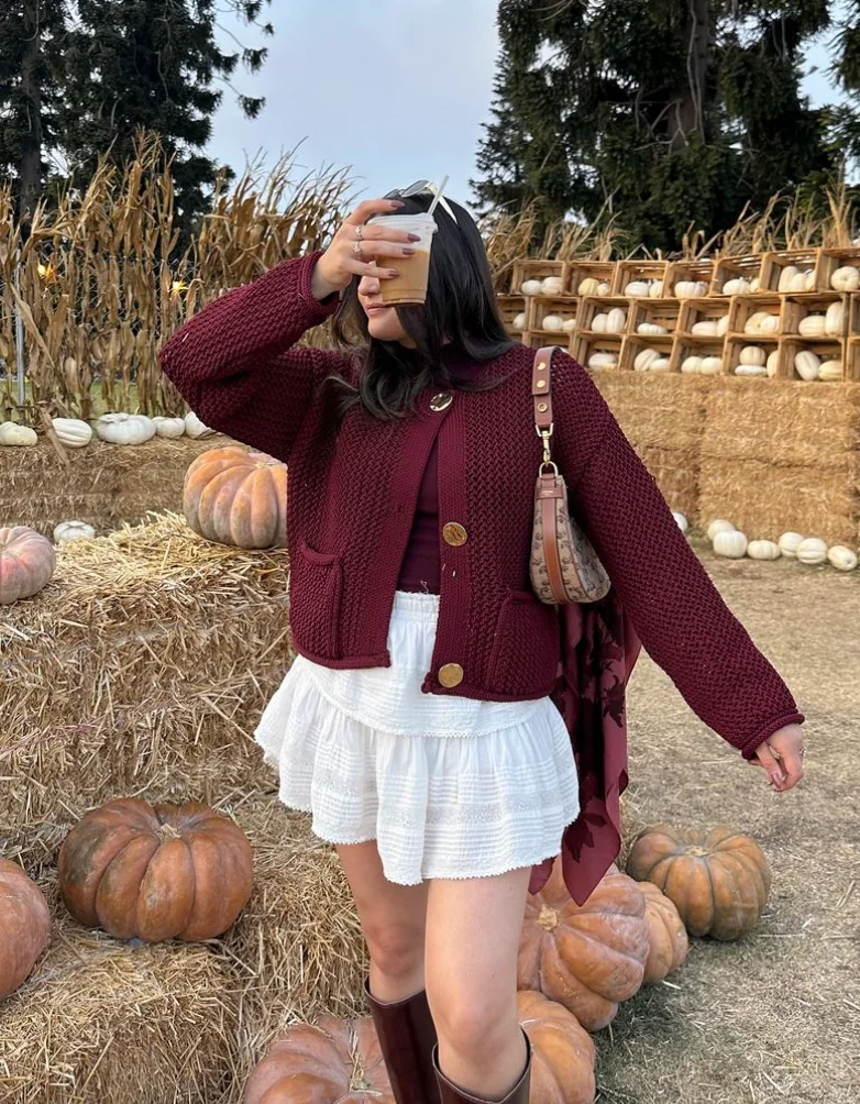 Woman in a burgundy outfit featuring a chunky knit cardigan over a burgundy top, styled with a white tiered mini skirt and brown knee-high boots, holding a drink at a pumpkin patch.