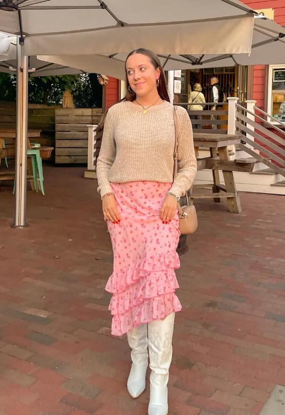 Early Fall Outfit featuring a cozy beige sweater tucked into a pink ruffled floral midi skirt, paired with tall white cowboy boots for a soft and feminine seasonal look.