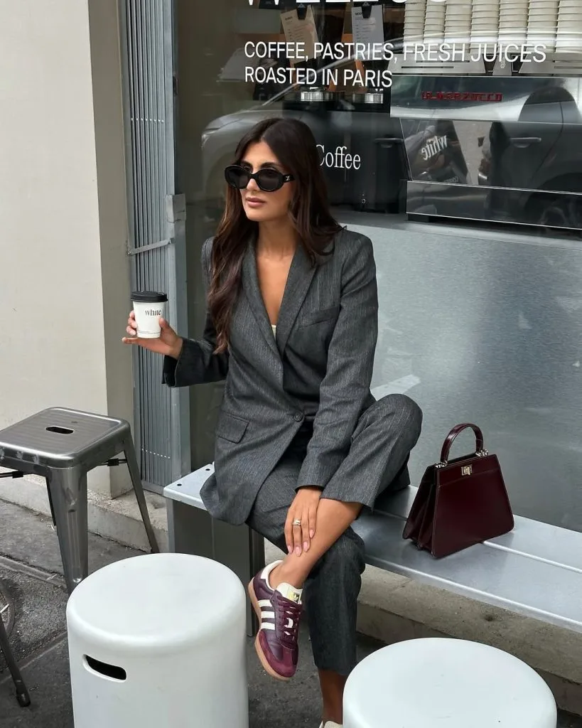 Office outfit in a tailored dark grey suit with a single-breasted blazer and matching trousers, styled casually with burgundy Adidas sneakers and a structured burgundy handbag.