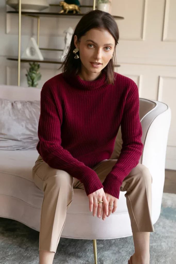 Woman seated indoors wearing a burgundy outfit with a ribbed turtleneck sweater and beige trousers, accessorized with statement earrings.